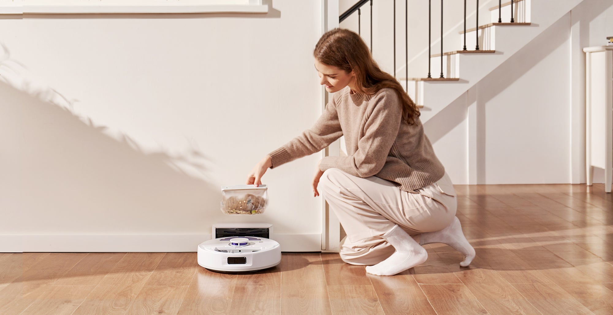 A woman placing a dustbin into the base station of the Narwal Freo X Plus Robot Vacuum and Mop, which is currently cleaning the floor.